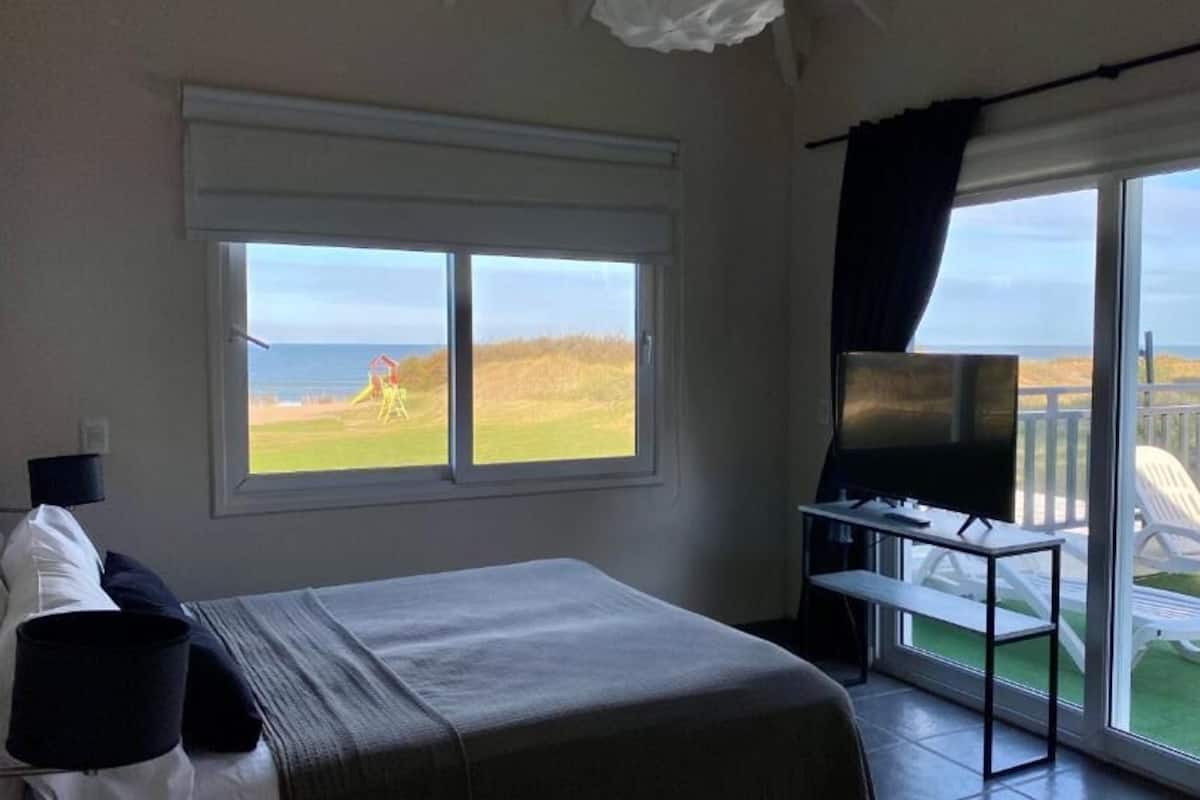 A bedroom with a bed, a TV, and a balcony with a view of the sea and grass.