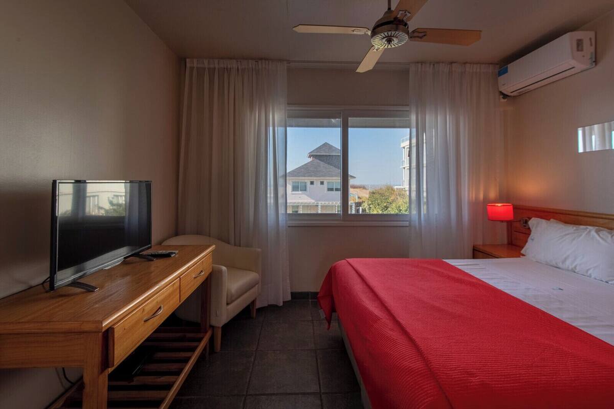A hotel room with a bed, a desk with a TV, a chair, a window with curtains, and a ceiling fan.