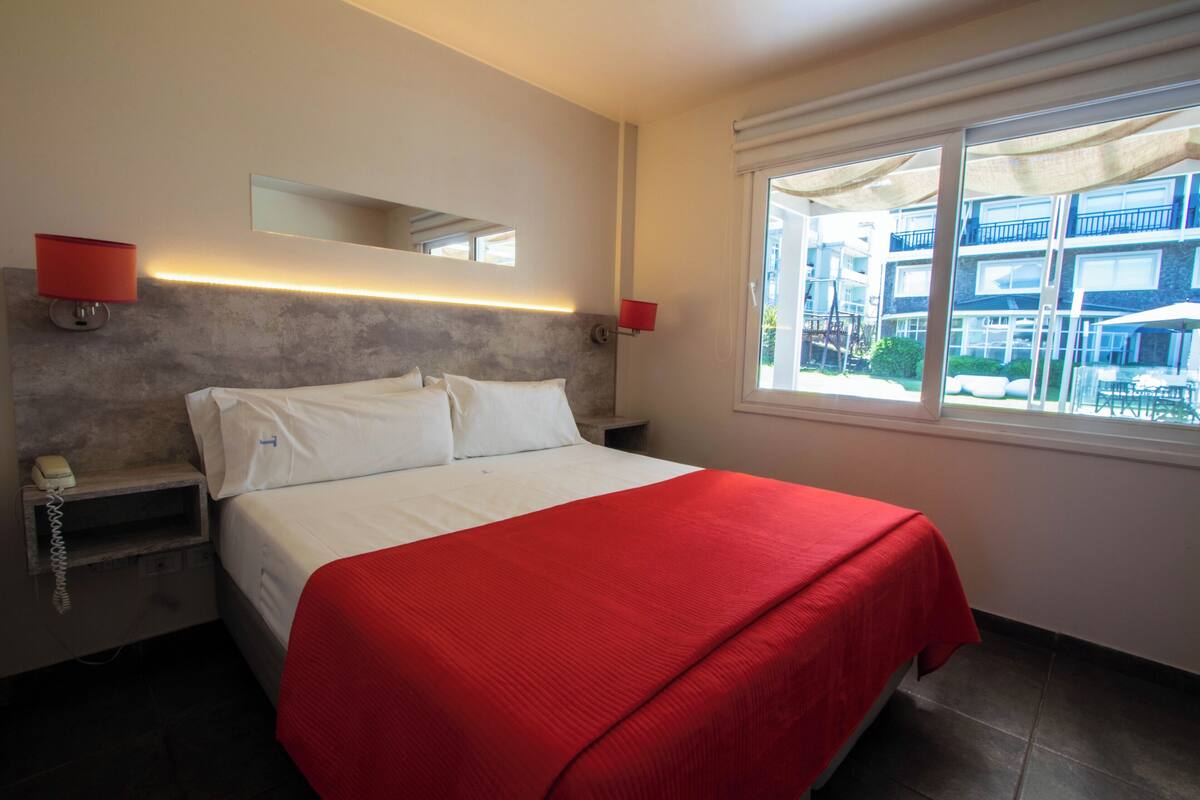 A hotel room with a large bed, a nightstand with a phone, a wall-mounted mirror, and a window with a view of the building's exterior.