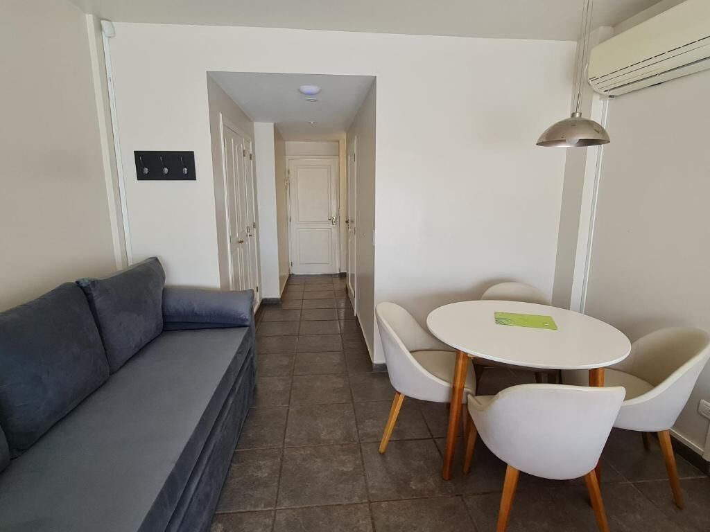 Un petit salon moderne comprenant un canapé, une table ronde et deux chaises. Une porte donne accès à une autre pièce, et un luminaire est fixé au mur.