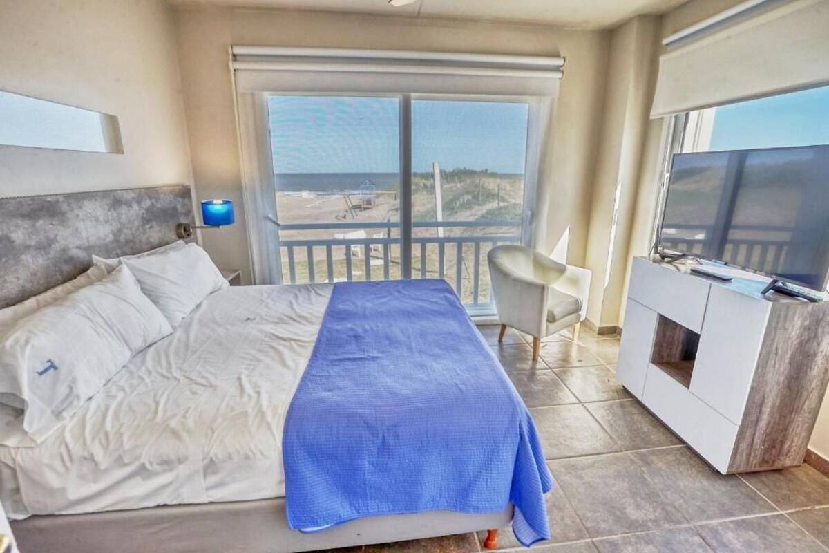 A bedroom with a bed, a TV, a desk, and a balcony with a view of the beach.