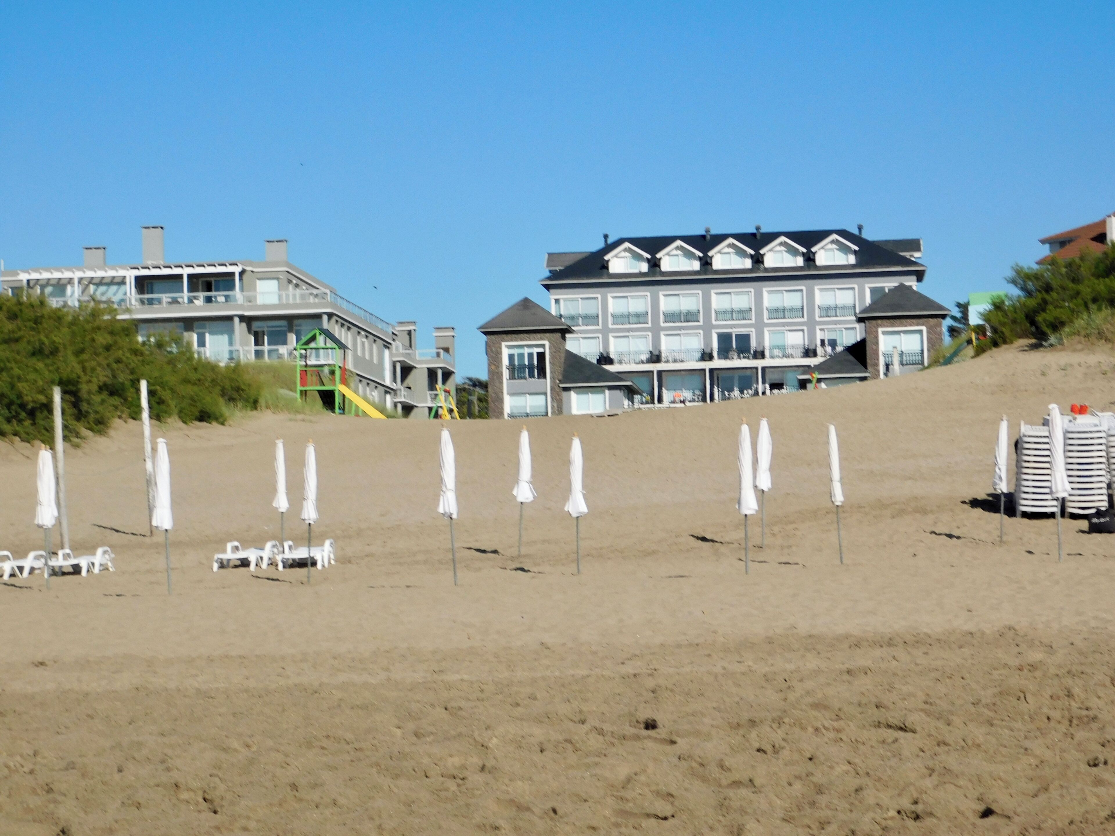 Une plage avec des parasols blancs et des chaises longues, un grand bâtiment doté de nombreuses fenêtres, et un escalier menant à celui-ci.