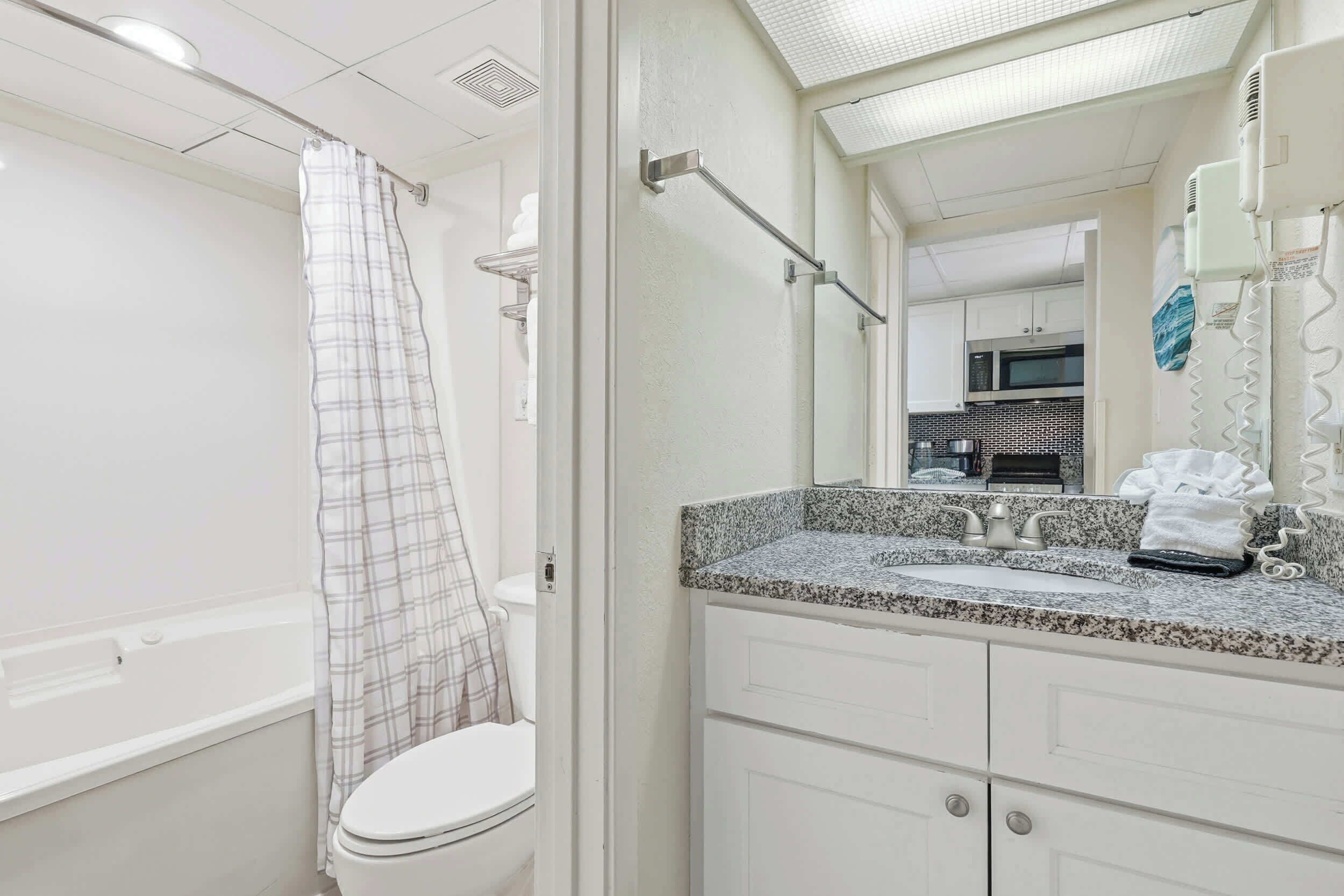 A bathroom with a white toilet, a shower with a plaid curtain, a granite countertop sink, and a mirror reflecting a kitchen area.