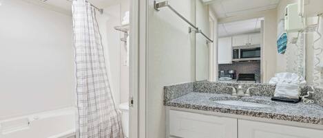 A bathroom with a white toilet, a shower with a plaid curtain, a granite countertop sink, and a mirror reflecting a kitchen area.