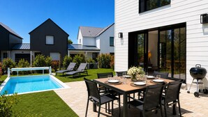 Un área de comedor moderna al aire libre con piscina, sillas de descanso y una parrilla.