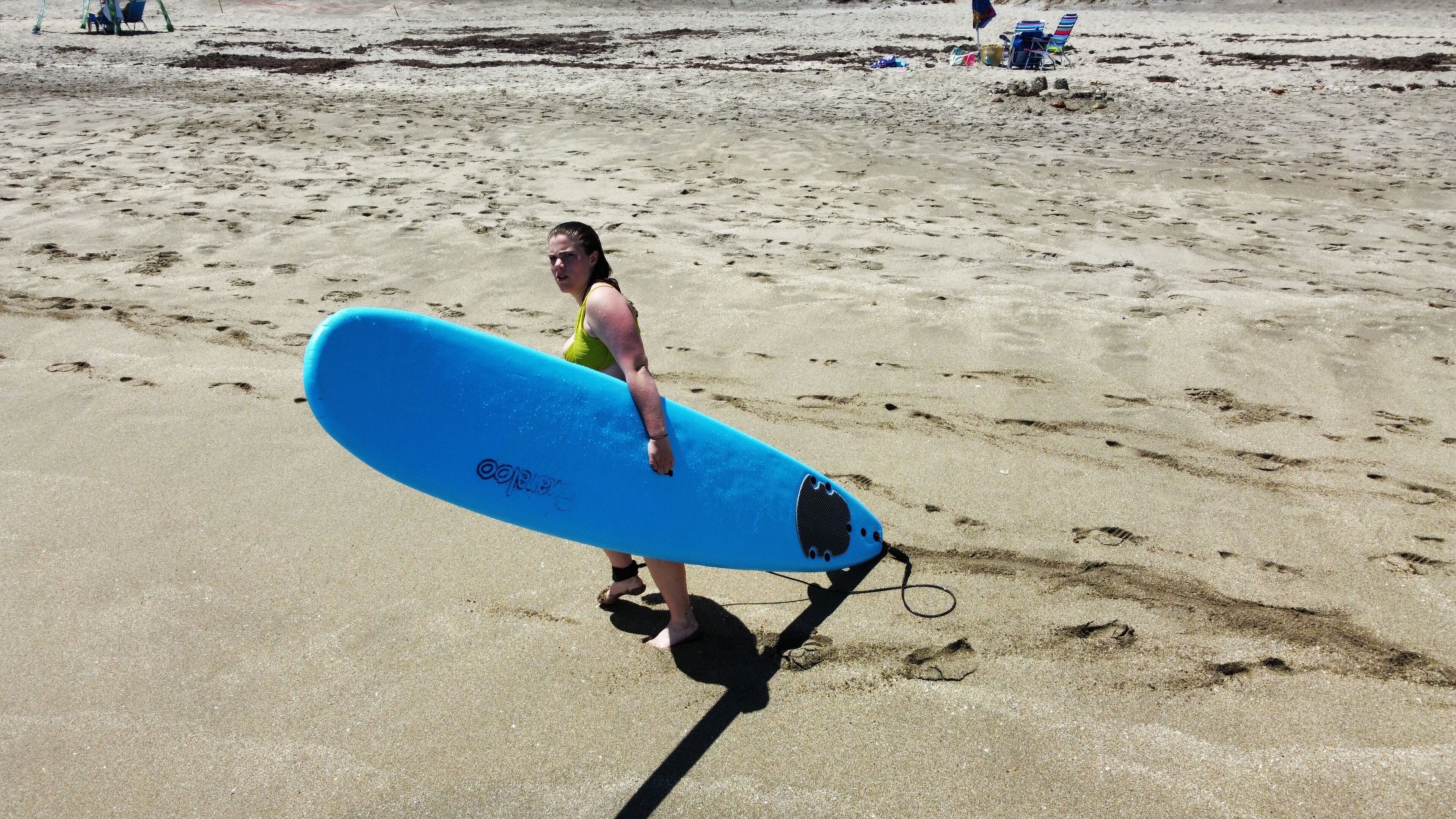 A person holding a blue surfboard on a sandy beach with footprints and beach chairs in the background.