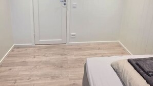 Ett sovrum med ett trägolv, en säng med en vit madrass och en grå filt, en vit dörr med ett handtag och en liten trähylla på väggen.