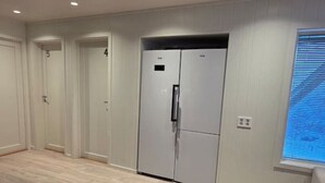 Una habitación amplia con un refrigerador blanco grande, pisos de madera y una ventana con persianas.