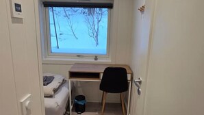 Un pasillo estrecho que conduce a un dormitorio con una cama, un escritorio y un ventanal con vista a árboles.