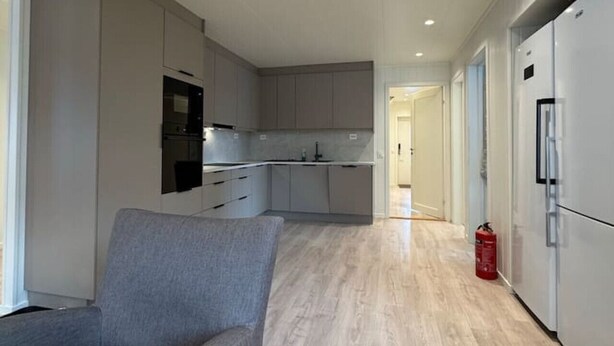 Una cocina moderna con armarios grises, horno integrado y microondas. Hay un sofá gris y una pequeña mesa en la sala de estar.