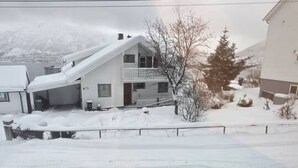 Una casa de dos pisos con garaje cubierto, rodeada de terreno y árboles nevados.