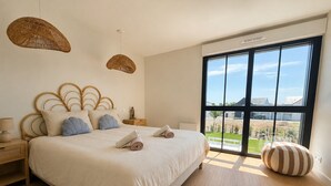 Une chambre moderne avec un grand lit, des tables de chevet, une lampe suspendue et une vue dégagée sur l’extérieur grâce à une grande fenêtre.