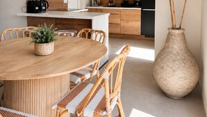 Une cuisine moderne avec des meubles en bois, un coin repas avec une table ronde et des chaises en osier, et un grand vase contenant des plantes séchées.