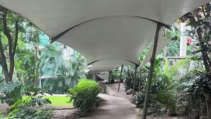 Un chemin de béton bordé d’une végétation luxuriante et de rochers, et surmonté d’une pergola blanche.