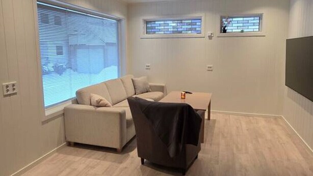 Un salon moderne avec un canapé beige, un fauteuil noir et un plancher en bois. Il y a deux fenêtres équipées de stores et un téléviseur à écran plat fixé au mur.