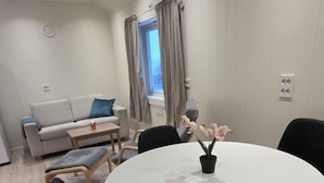 Un coin repas avec une table ronde, des chaises noires et une orchidée en pot. On aperçoit un canapé et une fenêtre avec des rideaux au fond.