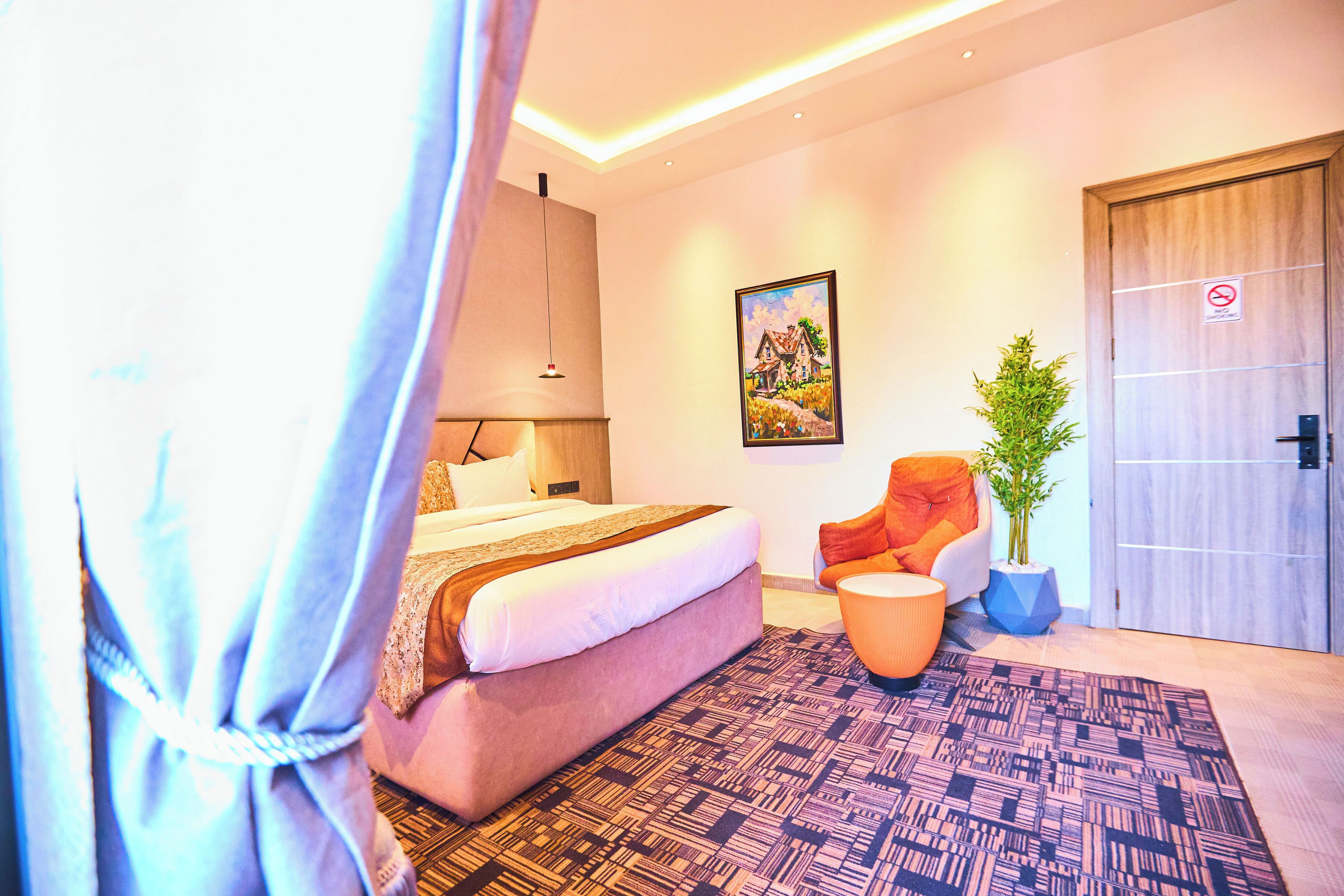 A modern hotel room with a bed, a chair, a potted plant, and a painting on the wall.