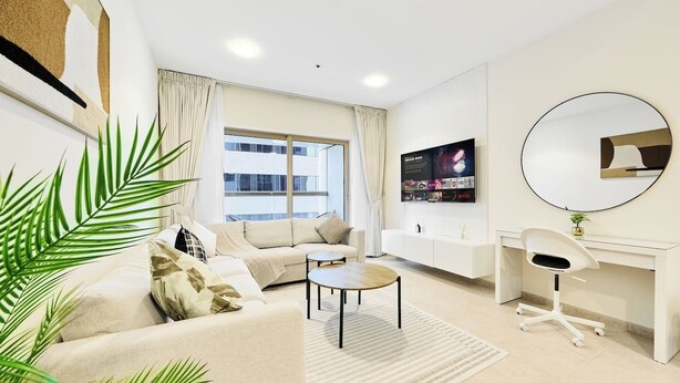 Uma sala de estar moderna com sofá, mesa de centro e uma escrivaninha com cadeira. Há uma janela grande com cortinas, uma televisão de tela plana na parede e um espelho redondo sobre a escrivaninha.