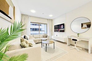 En moderne stue med sofa, sofabord og et skrivebord med stol. Der er et stort vindue med gardiner, et fladskærms-tv monteret på væggen og et rundt spejl over skrivebordet.