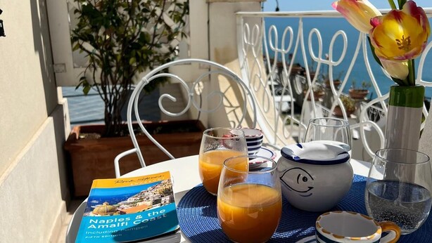 Una mesa lista para el desayuno, con un libro, vasos de zumo de naranja, una tetera y una taza, en una terraza con vistas al mar.