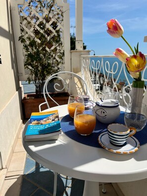 Una mesa lista para el desayuno, con un libro, vasos de zumo de naranja, una tetera y una taza, en una terraza con vistas al mar.