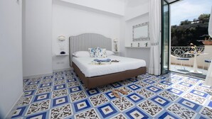 Un dormitorio con cabecera a rayas, cama con ropa de cama blanca, suelo con estampado azul y blanco, balcón con barandilla blanca y vistas a vegetación y un puente.