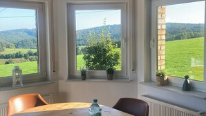 A dining area with a round wooden table, three chairs, a bottle of water, and a box of tissues. There are three windows with views of green fields and hills. - Country life, quiet and modern on our farm, bay window apartment with great views (Windeck)