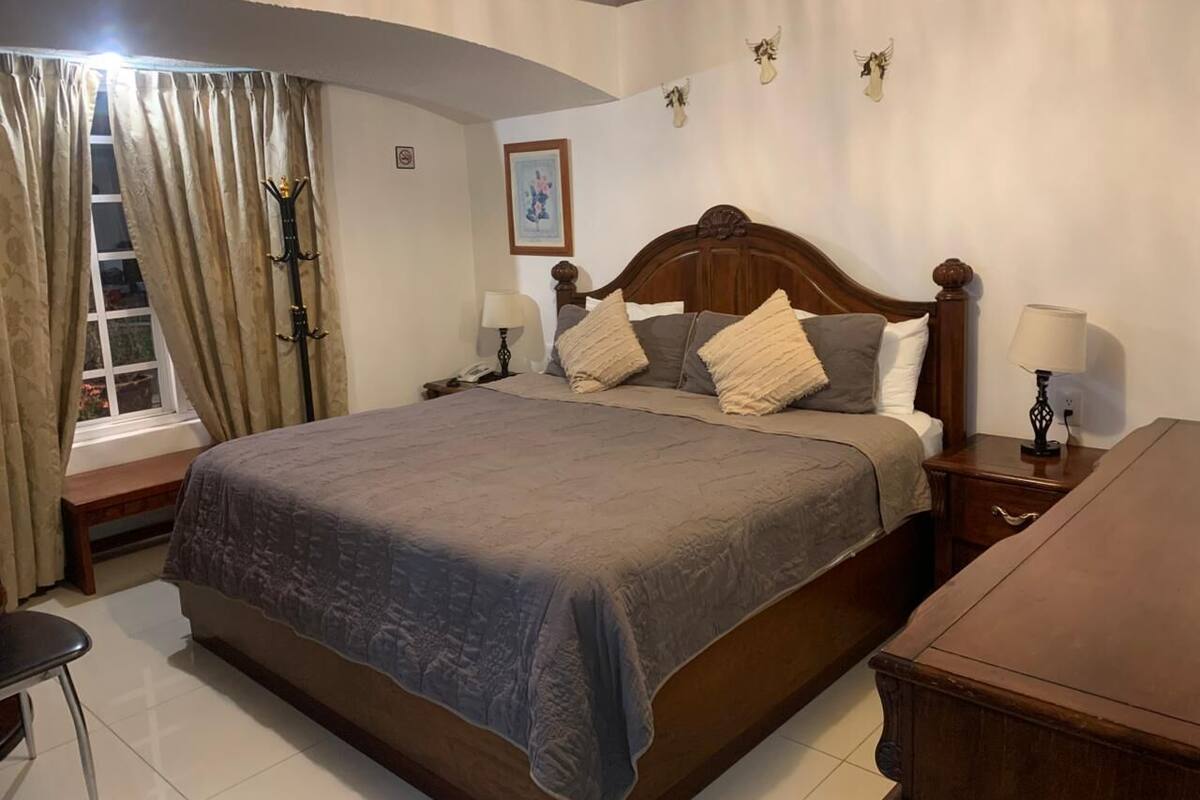 Un dormitorio ordenado con una cama grande, mesitas de noche, un banco de madera y una ventana con cortinas.