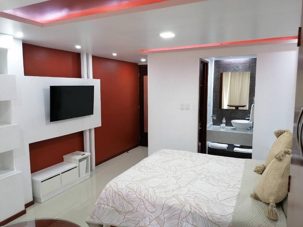 Un dormitorio moderno con cama, televisor montado en la pared, espejo y baño visible a través de una puerta abierta.