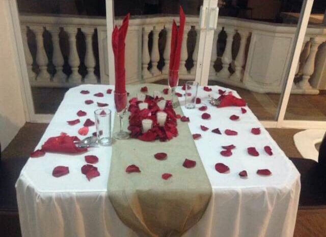 A table set with red rose petals, a candle, and a glass of water.