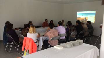 Un salón de conferencias con pantalla de presentación, mesas, sillas y una mesa con platos y servilletas.