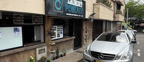 A building with a laundry service sign, a Mercedes-Benz parked in front, and potted plants on the sidewalk.