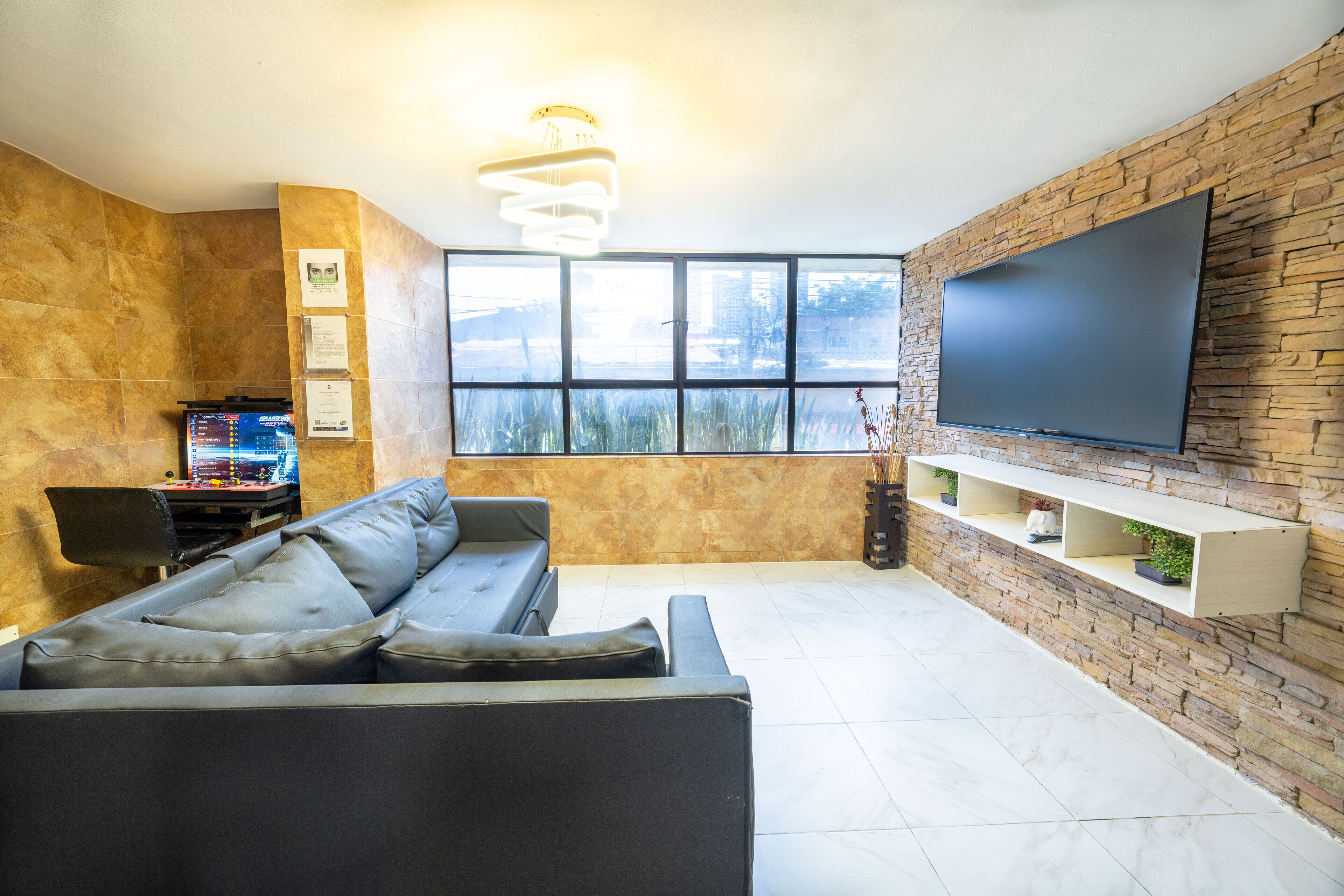 A modern living room with a sectional sofa, a flat-screen TV mounted on a stone wall, and a large window providing a view of the outdoors.