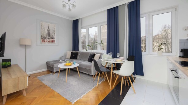 A modern living room with a sofa, dining table, and chairs, a small kitchen area, and large windows with curtains. - Apartment with Balcony in the Center | Air Conditioning and View (Warszawa)