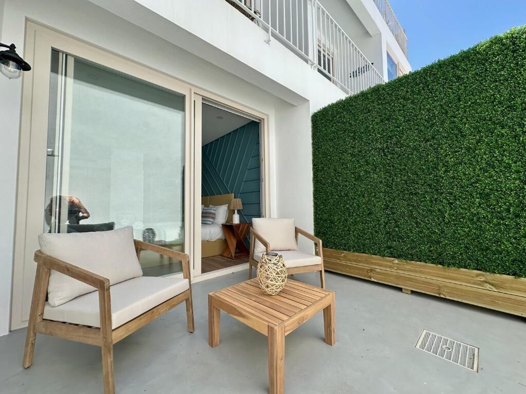 A modern outdoor seating area with two wooden chairs and a table, a glass door leading inside, and a green hedge wall.