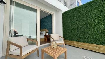 A modern outdoor seating area with two wooden chairs and a table, a glass door leading inside, and a green hedge wall.