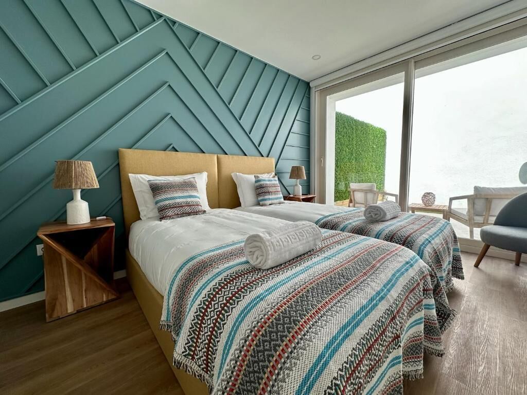 A modern hotel room with a large bed, two bedside tables, and a view of a green hedge through the window.