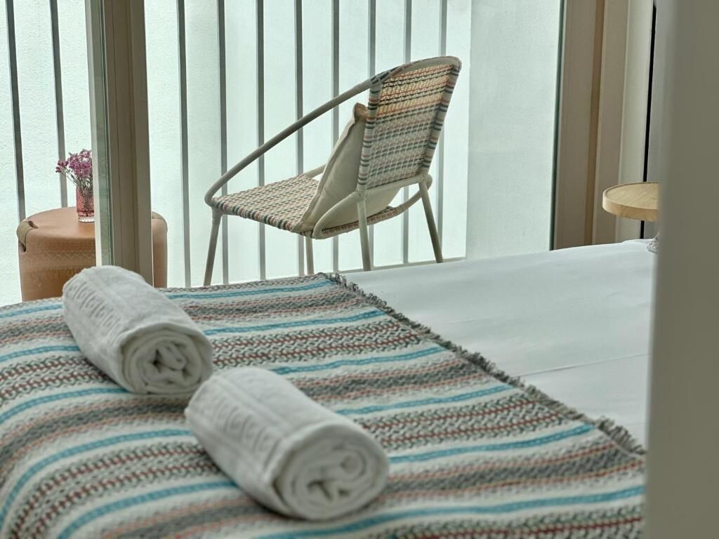 Una camera d'albergo con un letto, un tappeto a righe, un asciugamano arrotolato, una sedia in vimini e un tavolino con un vaso.