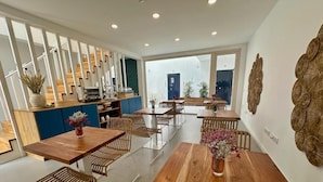 Un comedor moderno con mesas de madera y sillas de mimbre, una zona de cocina con armarios azules y detalles decorativos en la pared.