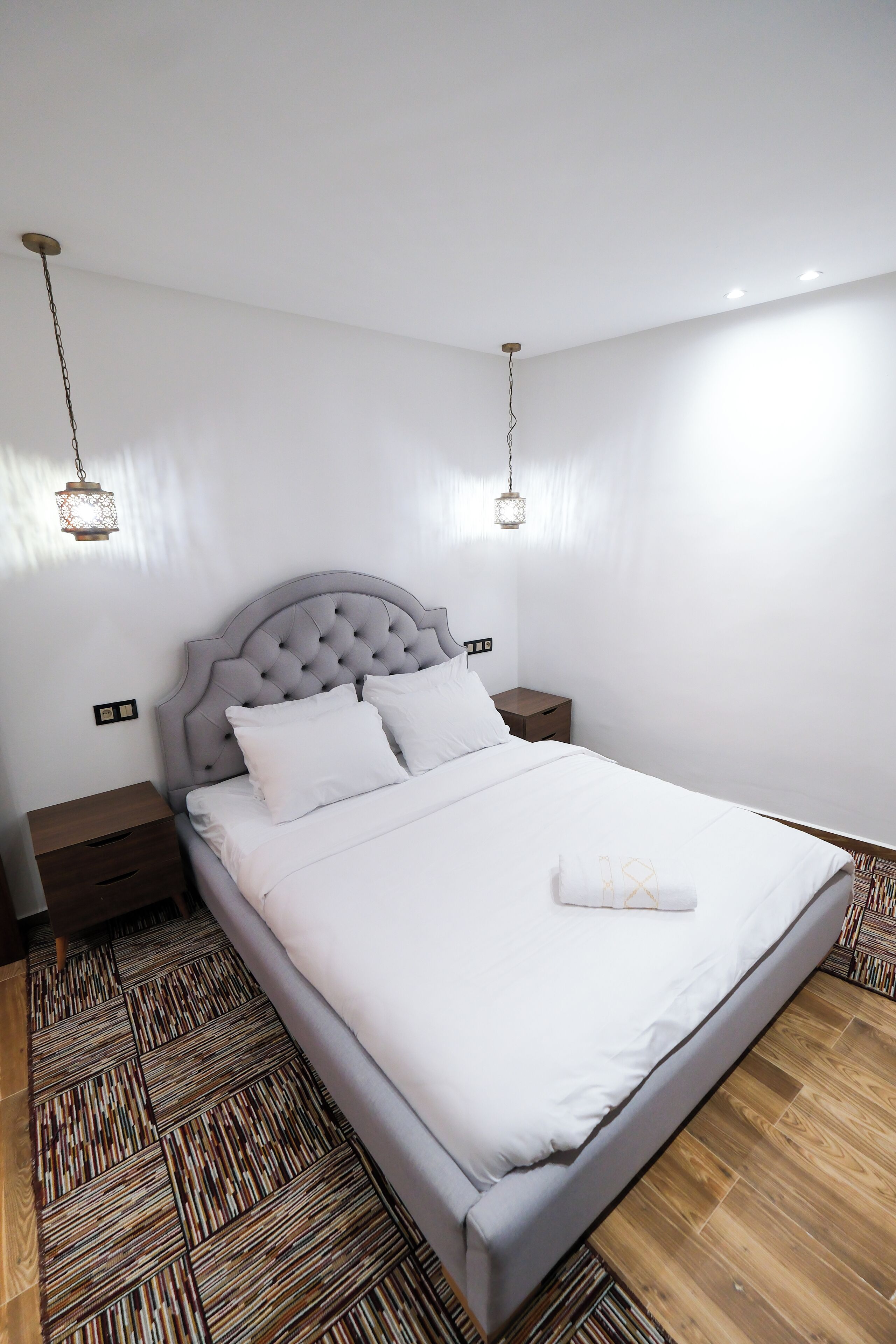Una cama bien hecha con sábanas blancas, un cabecero capitonado y una mesita de noche con una lámpara.