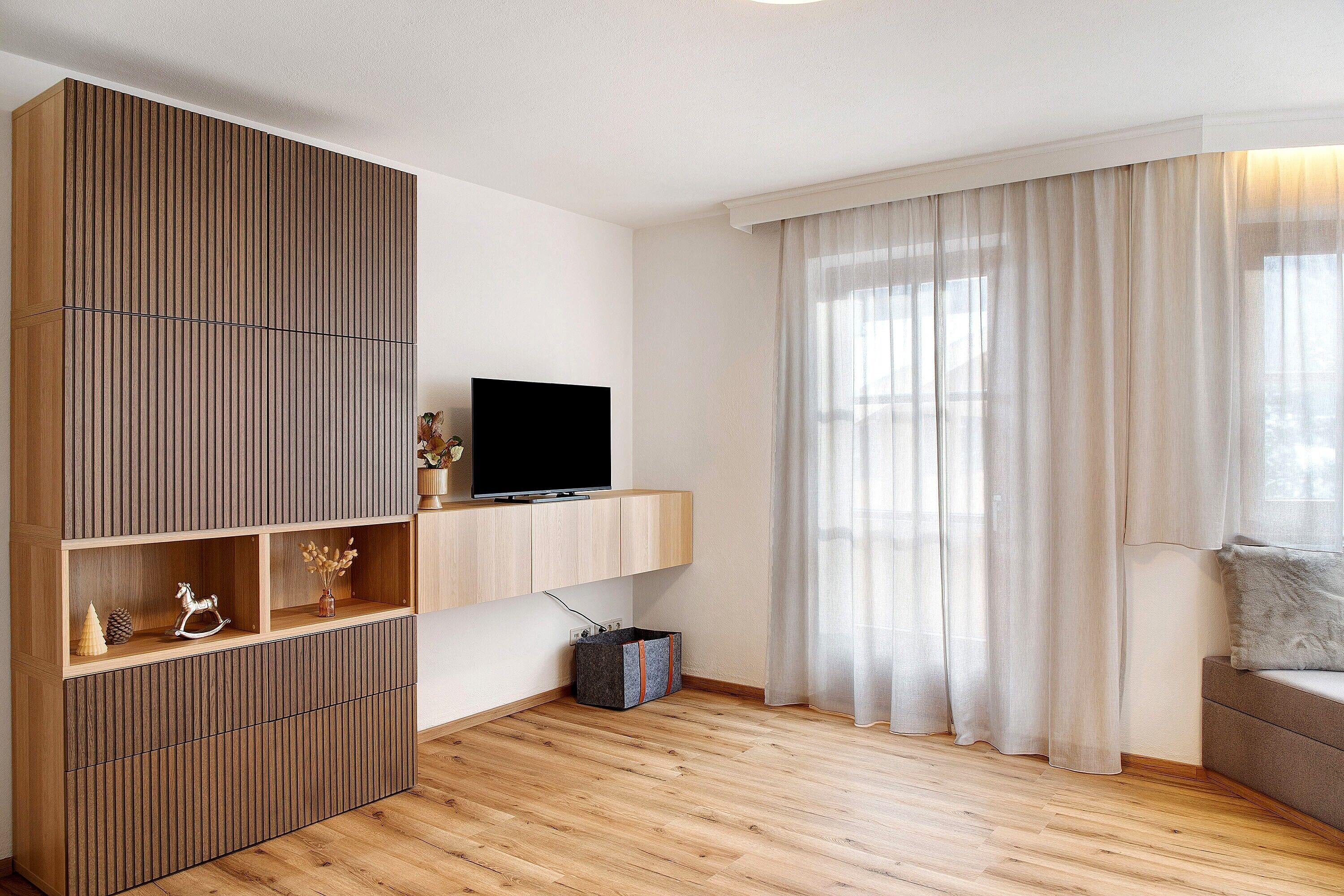 Ein modernes Wohnzimmer mit einer Holzeinrichtungsanlage, einem Flachbildfernseher, einer Couch und einem großen Fenster mit durchscheinenden Vorhängen.