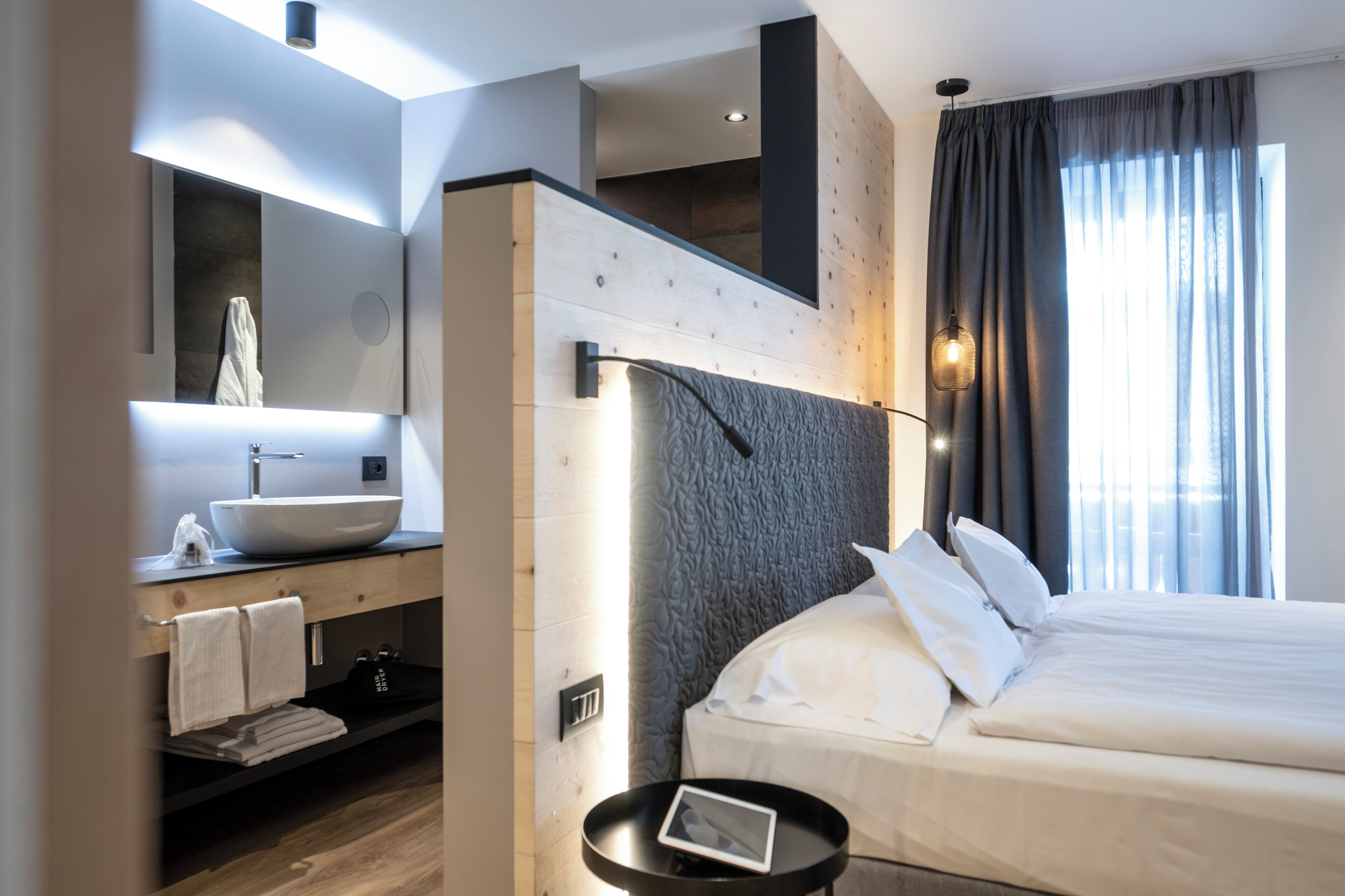 A modern hotel room with a large bed, a built-in vanity with a large mirror, a round bedside table, and a view of the outdoors through a window with curtains.