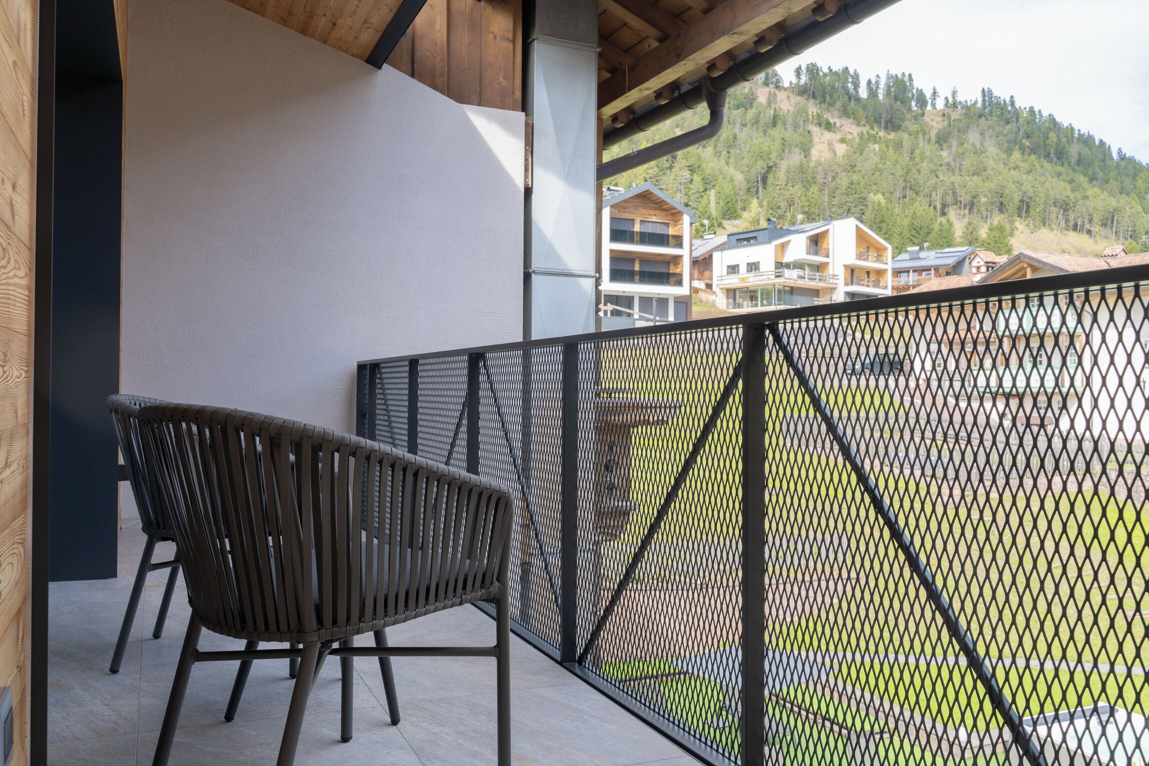 Un balcon doté d’une rambarde en métal, d’une chaise en bois et offrant une vue sur des immeubles et une colline boisée en arrière-plan.