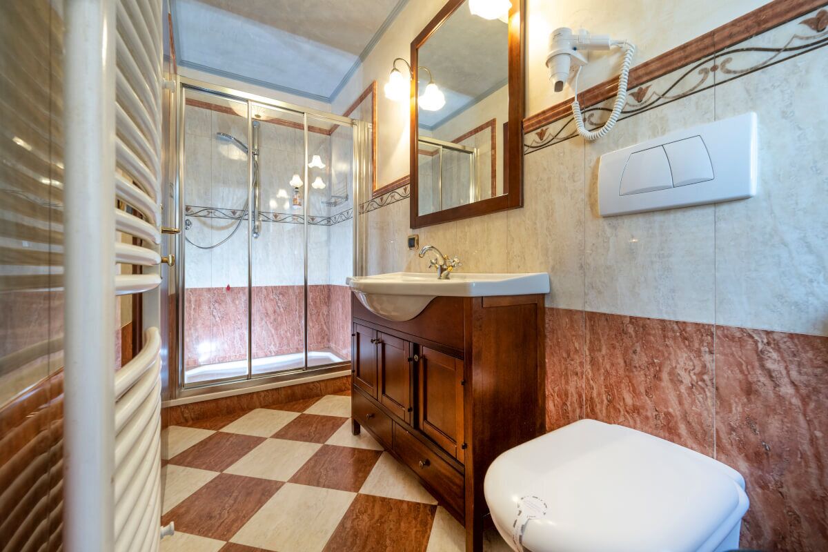 Ein Badezimmer mit Schachbrettboden, einem weißen Waschbecken, einer Holzkommode und einer Duschkabine aus Glas, sowie einem großen Spiegel.