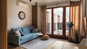 A modern living room with a blue sofa, a round wall clock, a small table with a vase, and a large window with curtains.
