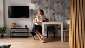 A modern living room with a flat-screen TV mounted on the wall, a dining table with chairs, a black metal shelf with decorative items, and a potted plant.