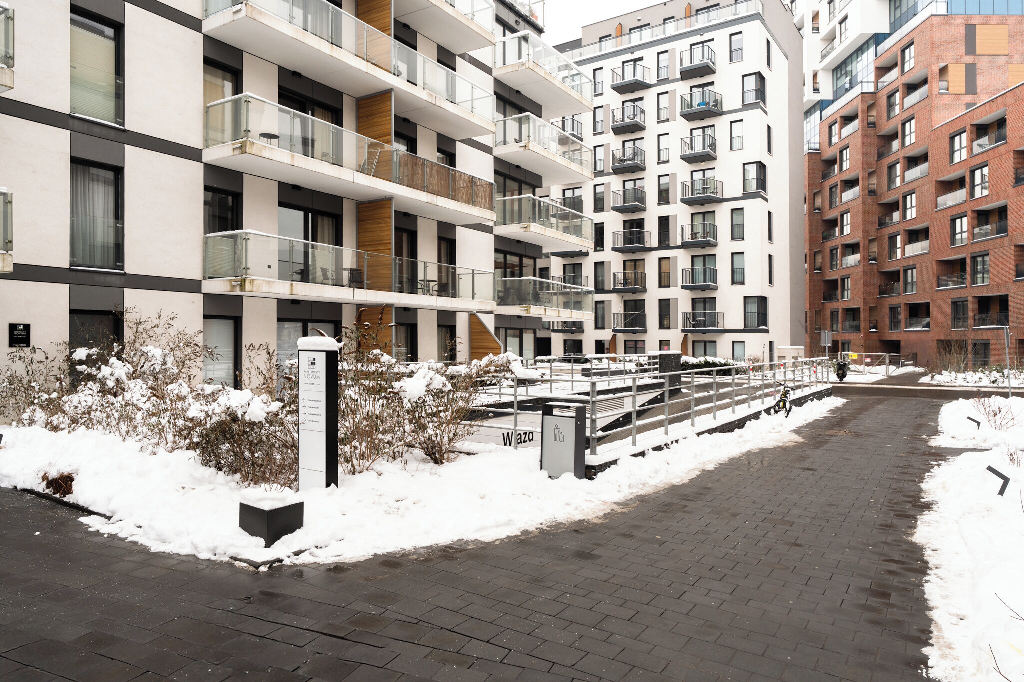 A multi-story residential building with balconies, a paved walkway, and a sign indicating a location.