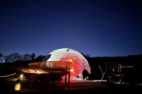 Another lit geodesic dome sits on a raised deck with outdoor seating and a fire pit under the night sky