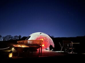 A dome-shaped structure with a fire in the center, surrounded by outdoor seating. - Cosy Off-Grid Stargazing Dome in the Jurassic Coast with optional Hot Tub & Sauna (Devon)