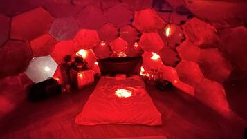 Una stanza con parete in pietra esagonale, un letto con coperta rossa e un lanterne appesa.